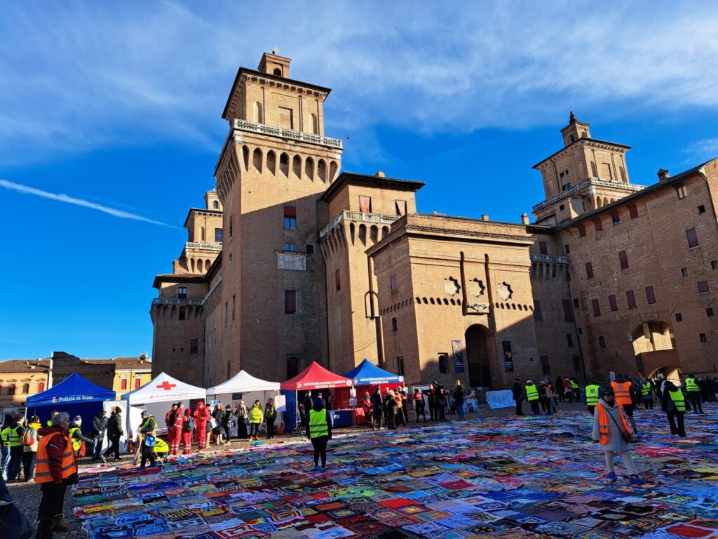 Foto del Castello Estense di Ferrara durante un evento pubblico, con stand di associazioni di volontariato come la Croce Rossa Italiana e Polizia di Stato. Il piazzale è decorato con una vasta esposizione di coperte colorate realizzate a mano, simbolo di solidarietà e creatività. Diversi volontari in divisa e visitatori partecipano alla manifestazione sotto un cielo azzurro. L’immagine cattura un momento di comunità e collaborazione in una delle piazze storiche della città.